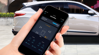 Closeup of hand holding a phone with AcuraLink app open on screen. In the background, a white Acura RDX parked in front of modern architectural building.